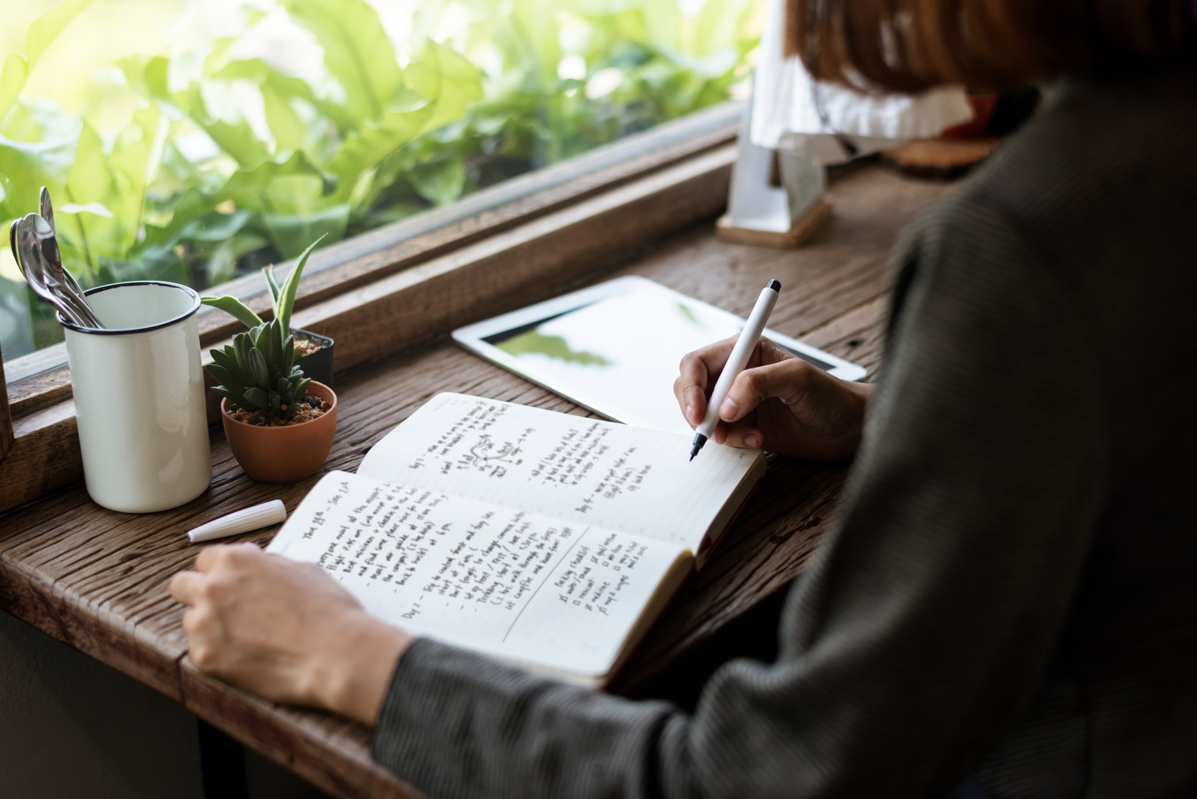Writer drafts memoir notes in notebook beside coffee and tablet.